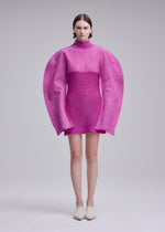 A person wears the ISSEY MIYAKE PANINI PULLED pink wool-alpaca blend dress with oversized, rounded sleeves and a high neck, standing against a plain light gray background.