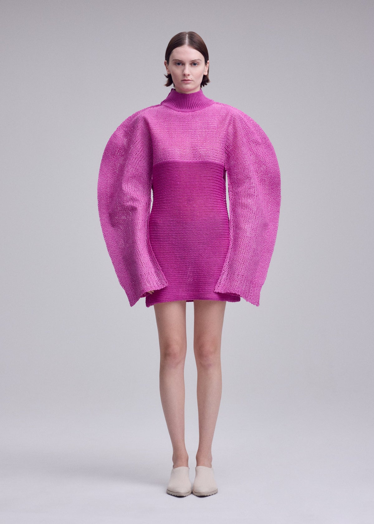 Zoomed view A person wears the ISSEY MIYAKE PANINI PULLED pink wool-alpaca blend dress with oversized, rounded sleeves and a high neck, standing against a plain light gray background.