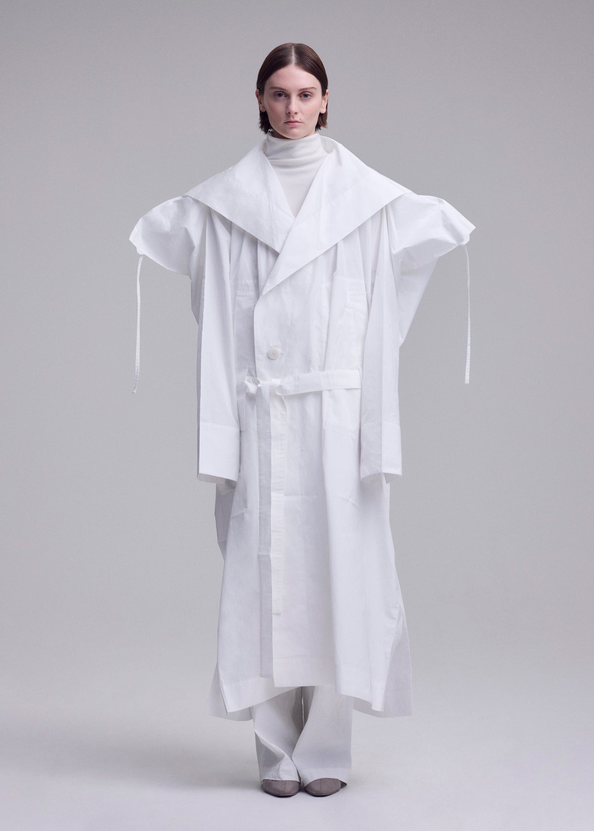 A person stands against a plain background wearing an oversized ISSEY MIYAKE FOLDING COAT with extended shoulders, a large collar, and long sleeves, paired with a white turtleneck and wide white pants. view 6 of 15