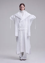A person stands against a plain background wearing an oversized ISSEY MIYAKE FOLDING COAT with extended shoulders, a large collar, and long sleeves, paired with a white turtleneck and wide white pants.