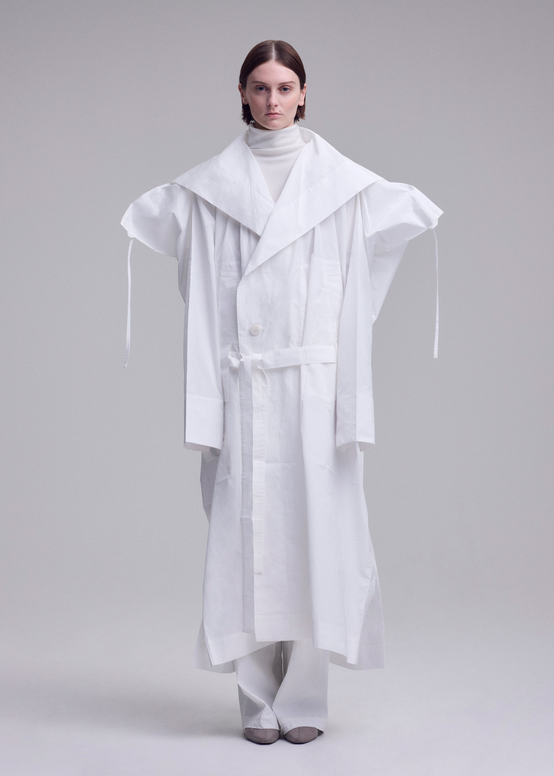 Zoomed view A person stands against a plain background wearing an oversized ISSEY MIYAKE FOLDING COAT with extended shoulders, a large collar, and long sleeves, paired with a white turtleneck and wide white pants.