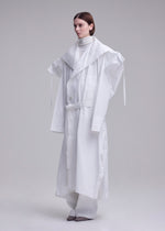 A person stands in a white studio wearing the ISSEY MIYAKE FOLDING COAT—an oversized, layered white coat with wide shoulders and a large collar—paired with loose white pants and gray shoes for an avant-garde, sculptural look.