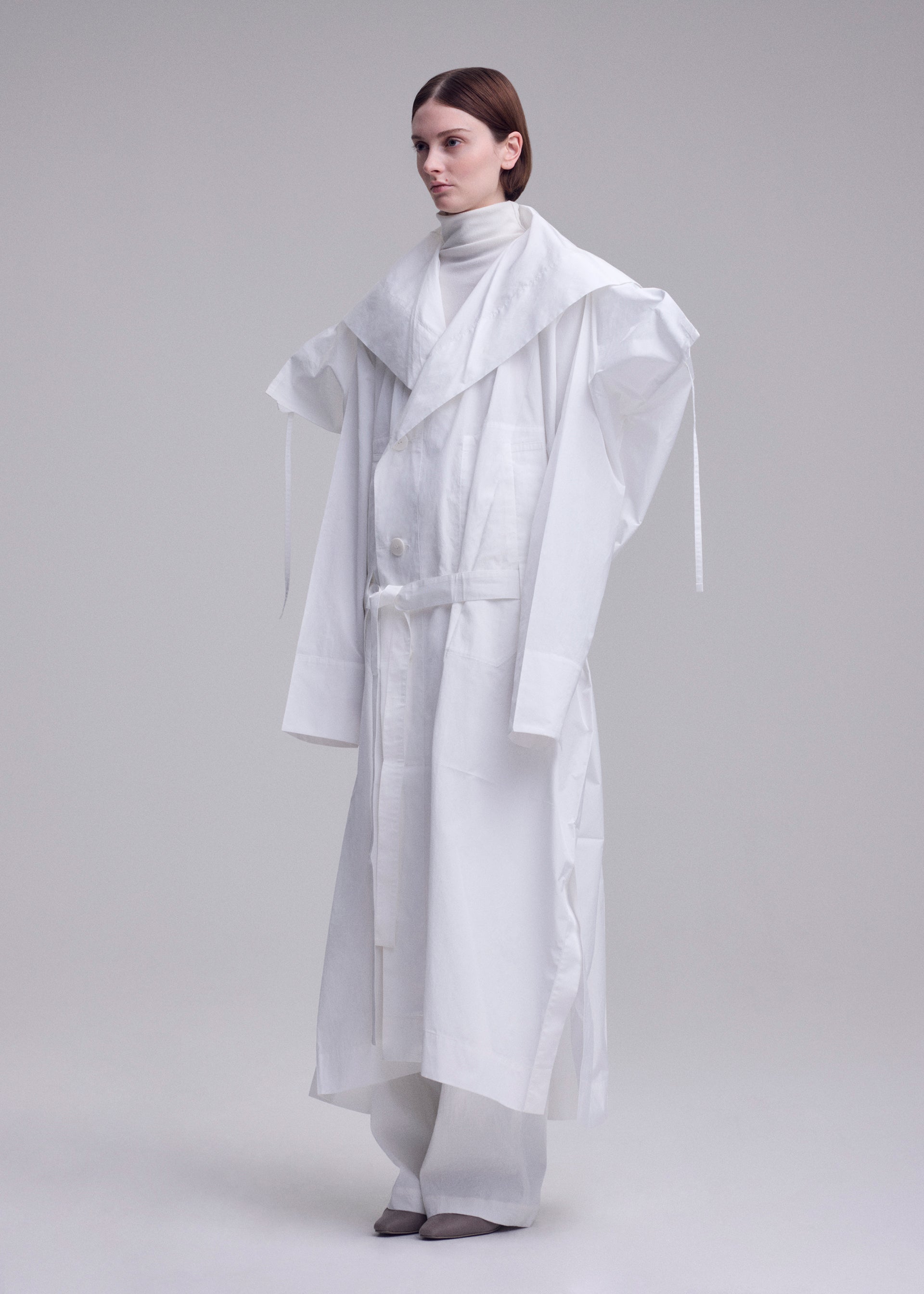Zoomed view A person stands in a white studio wearing the ISSEY MIYAKE FOLDING COAT—an oversized, layered white coat with wide shoulders and a large collar—paired with loose white pants and gray shoes for an avant-garde, sculptural look.