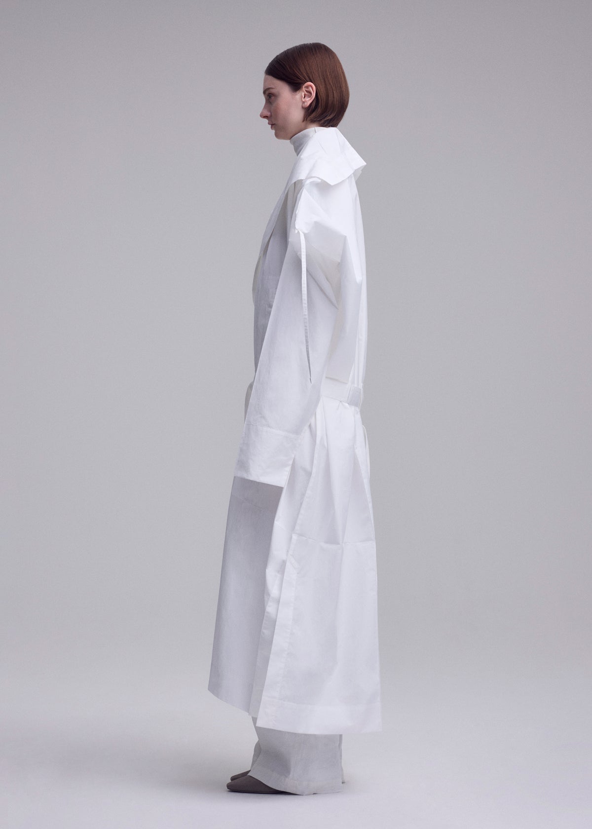 A person with short brown hair stands in profile wearing the ISSEY MIYAKE FOLDING COAT, featuring oversized sleeves and a high collar, set against a plain light gray background. view 9 of 15