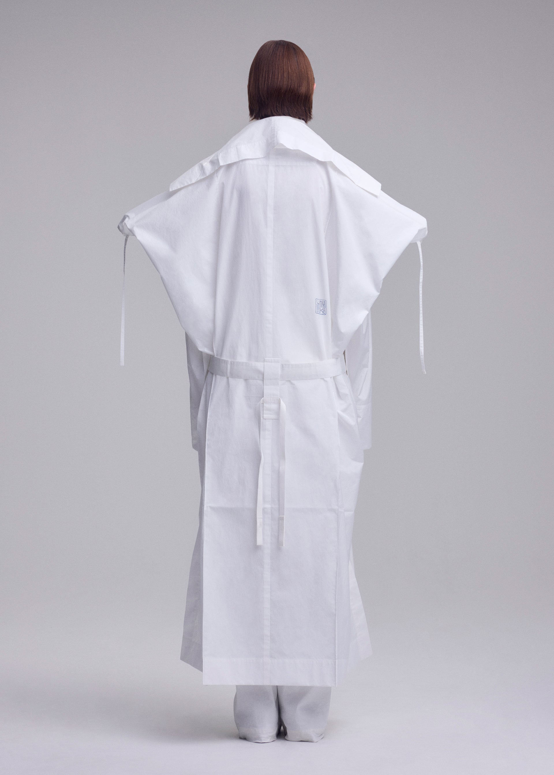Zoomed view A person stands facing away in ISSEY MIYAKE's FOLDING COAT, featuring a minimalist, structured design with wide sleeves and a belt, set against a plain light gray background.