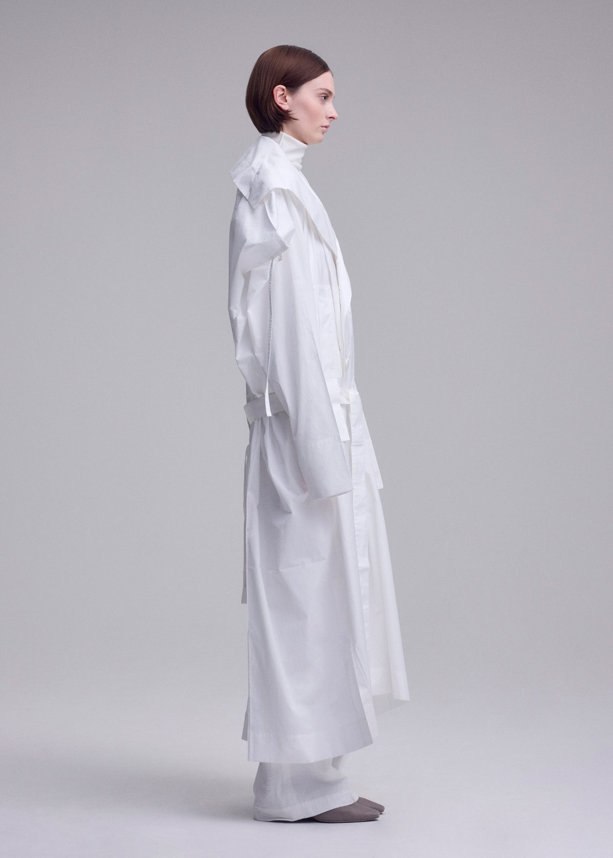 A person with short brown hair stands in profile against a light background, wearing ISSEY MIYAKE's long, loose-fitting FOLDING COAT with wide sleeves and white pants. Gray shoes are partially visible. view 10 of 15