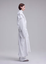 A person with short brown hair stands in profile against a light background, wearing ISSEY MIYAKE's long, loose-fitting FOLDING COAT with wide sleeves and white pants. Gray shoes are partially visible.