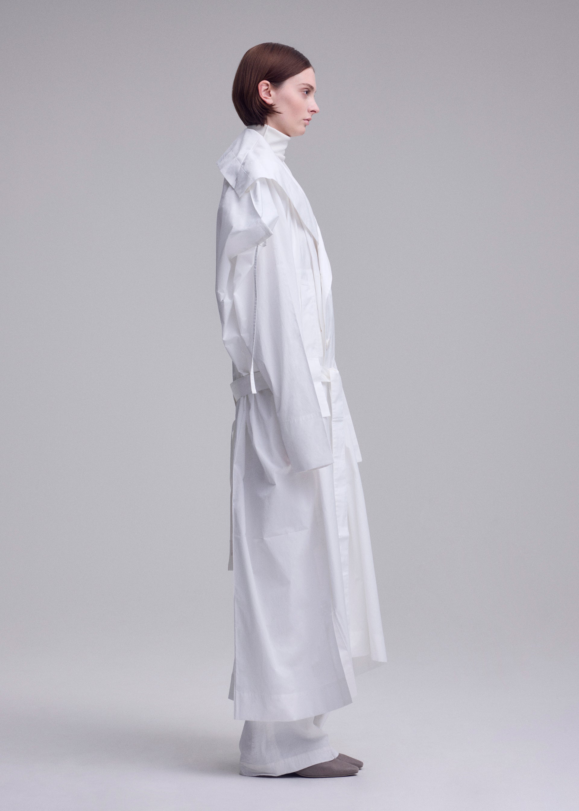 Zoomed view A person with short brown hair stands in profile against a light background, wearing ISSEY MIYAKE's long, loose-fitting FOLDING COAT with wide sleeves and white pants. Gray shoes are partially visible.