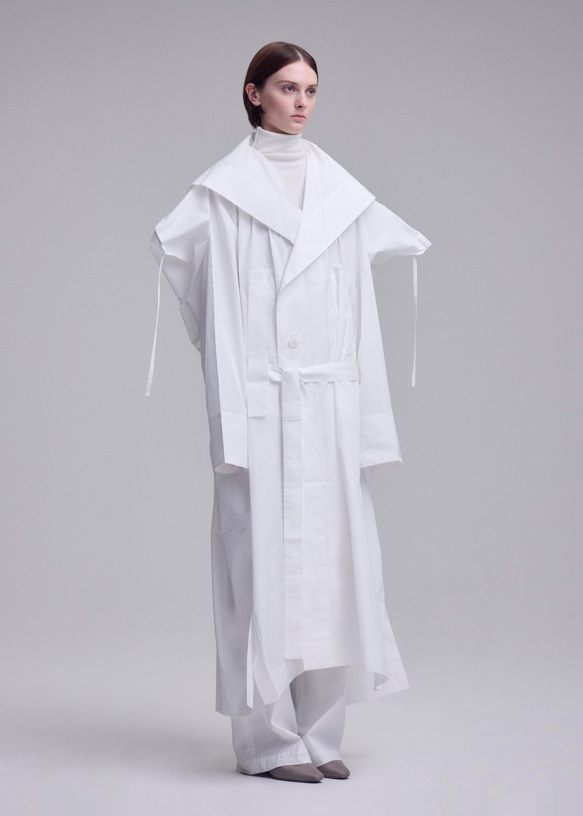 In a minimalist studio, a person wears ISSEY MIYAKE's oversized FOLDING COAT with wide lapels over a white turtleneck and loose pants. Their hair is sleek and straight, paired with grey pointed shoes. view 11 of 15