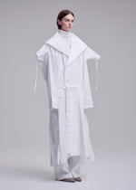 In a minimalist studio, a person wears ISSEY MIYAKE's oversized FOLDING COAT with wide lapels over a white turtleneck and loose pants. Their hair is sleek and straight, paired with grey pointed shoes.