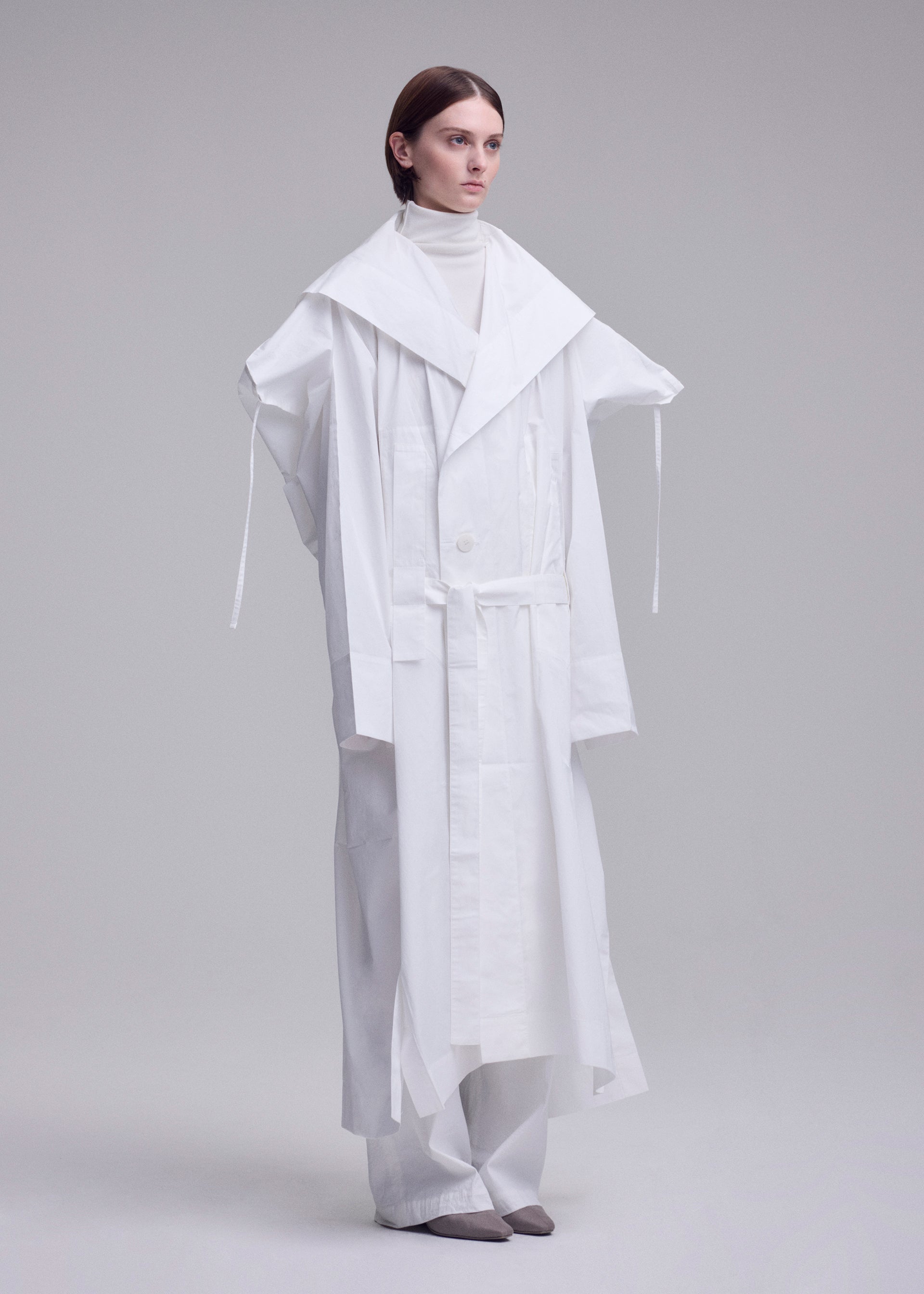 Zoomed view In a minimalist studio, a person wears ISSEY MIYAKE's oversized FOLDING COAT with wide lapels over a white turtleneck and loose pants. Their hair is sleek and straight, paired with grey pointed shoes.