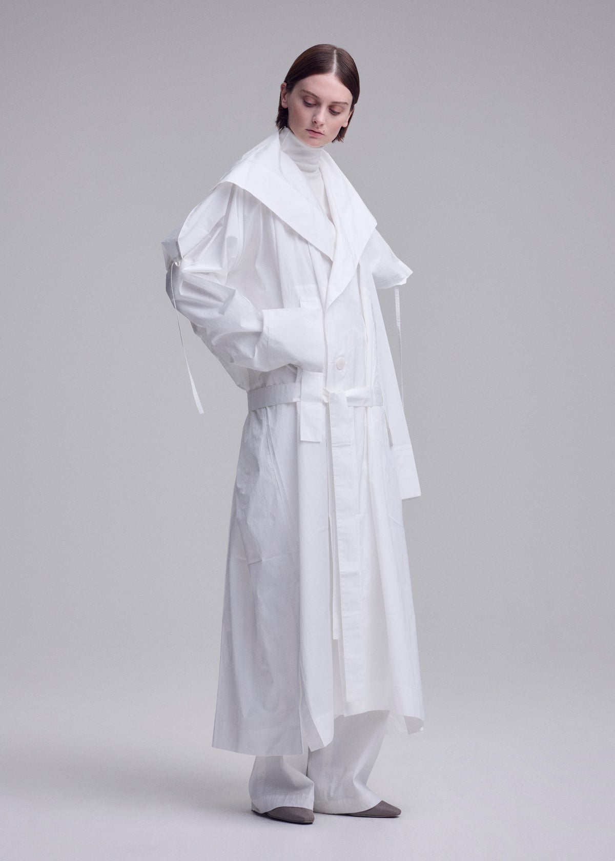 A person with short brown hair wears an ISSEY MIYAKE FOLDING COAT over white clothing and gray shoes, standing against a plain light gray background. view 15 of 15