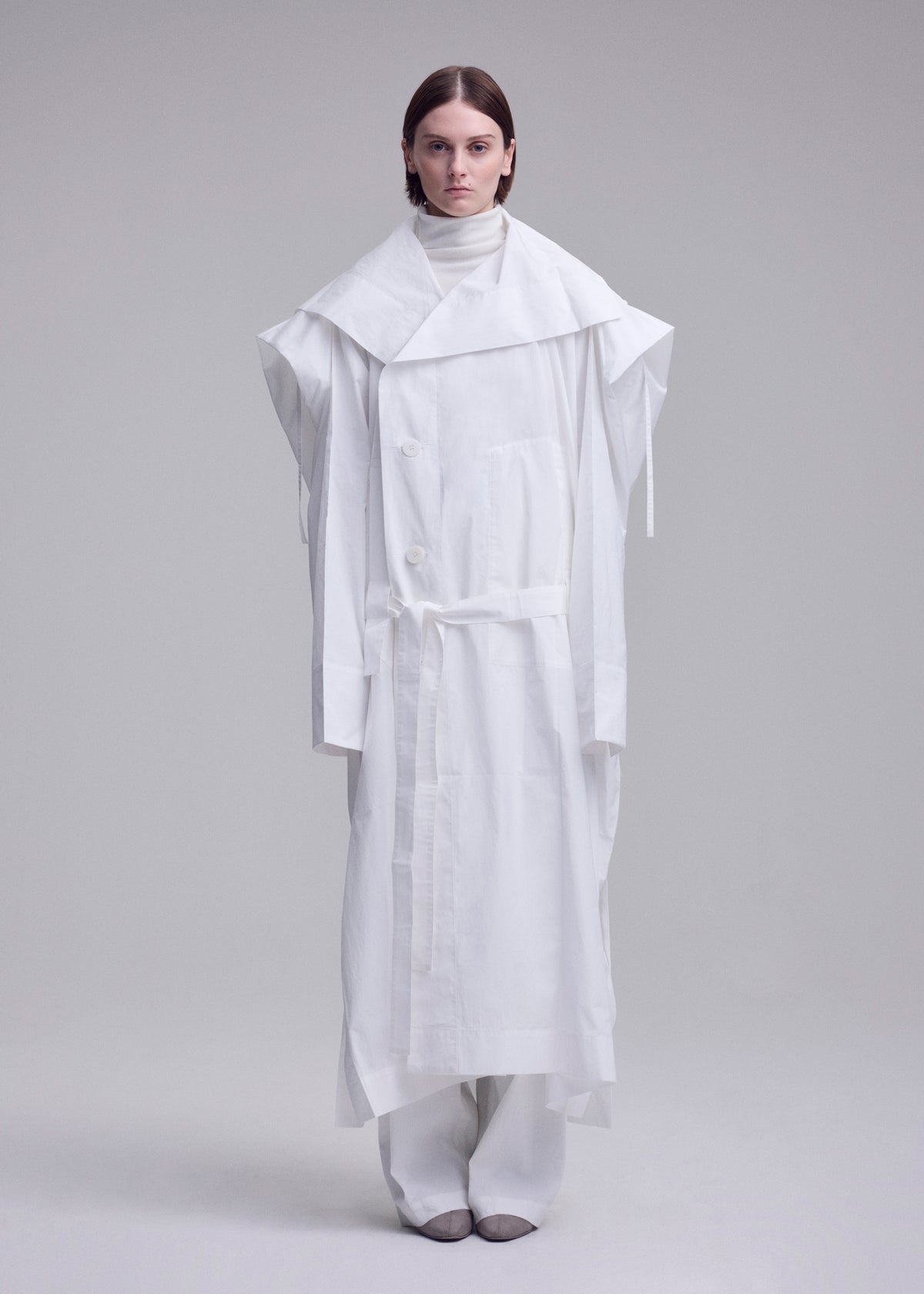 A person faces forward against a grey background, wearing the oversized ISSEY MIYAKE FOLDING COAT with wide shoulders and a belt, layered over white pants and a high-neck top. view 12 of 15