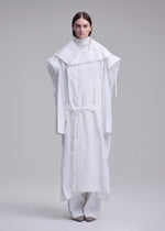 A person faces forward against a grey background, wearing the oversized ISSEY MIYAKE FOLDING COAT with wide shoulders and a belt, layered over white pants and a high-neck top.