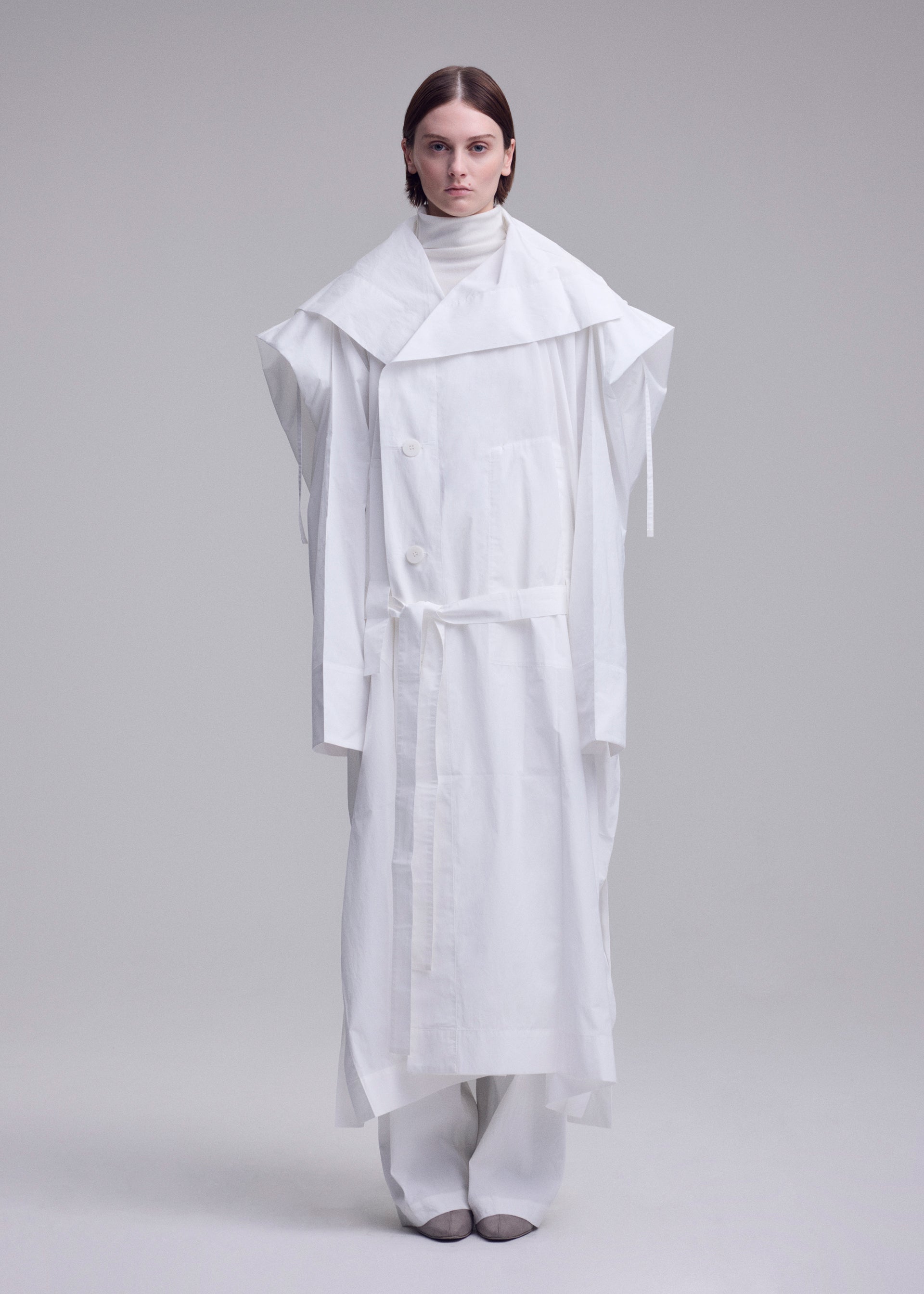 Zoomed view A person faces forward against a grey background, wearing the oversized ISSEY MIYAKE FOLDING COAT with wide shoulders and a belt, layered over white pants and a high-neck top.
