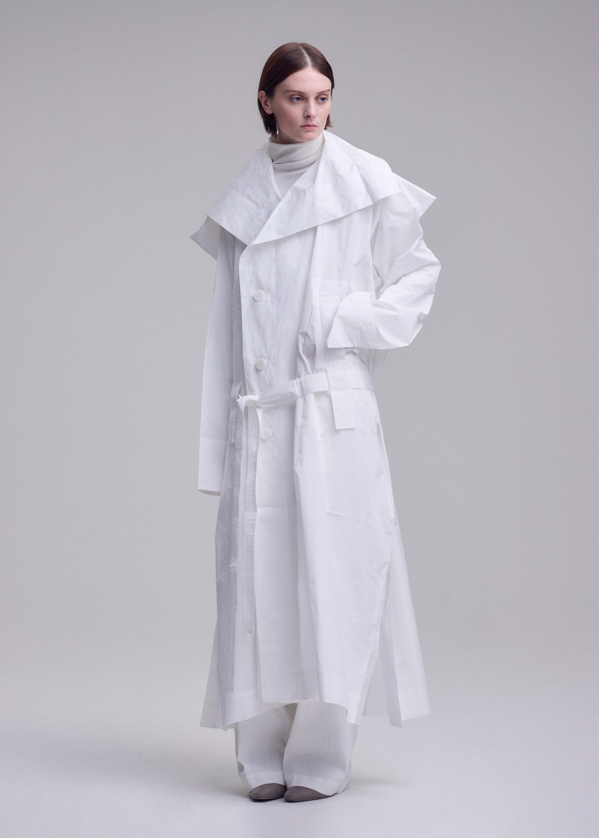 A person stands against a plain background wearing the ISSEY MIYAKE FOLDING COAT— a long white piece with a large collar, belted waist, and wide sleeves— layered over a high-neck top and wide-legged pants. view 14 of 15