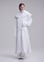 A person stands against a plain background wearing the ISSEY MIYAKE FOLDING COAT— a long white piece with a large collar, belted waist, and wide sleeves— layered over a high-neck top and wide-legged pants.