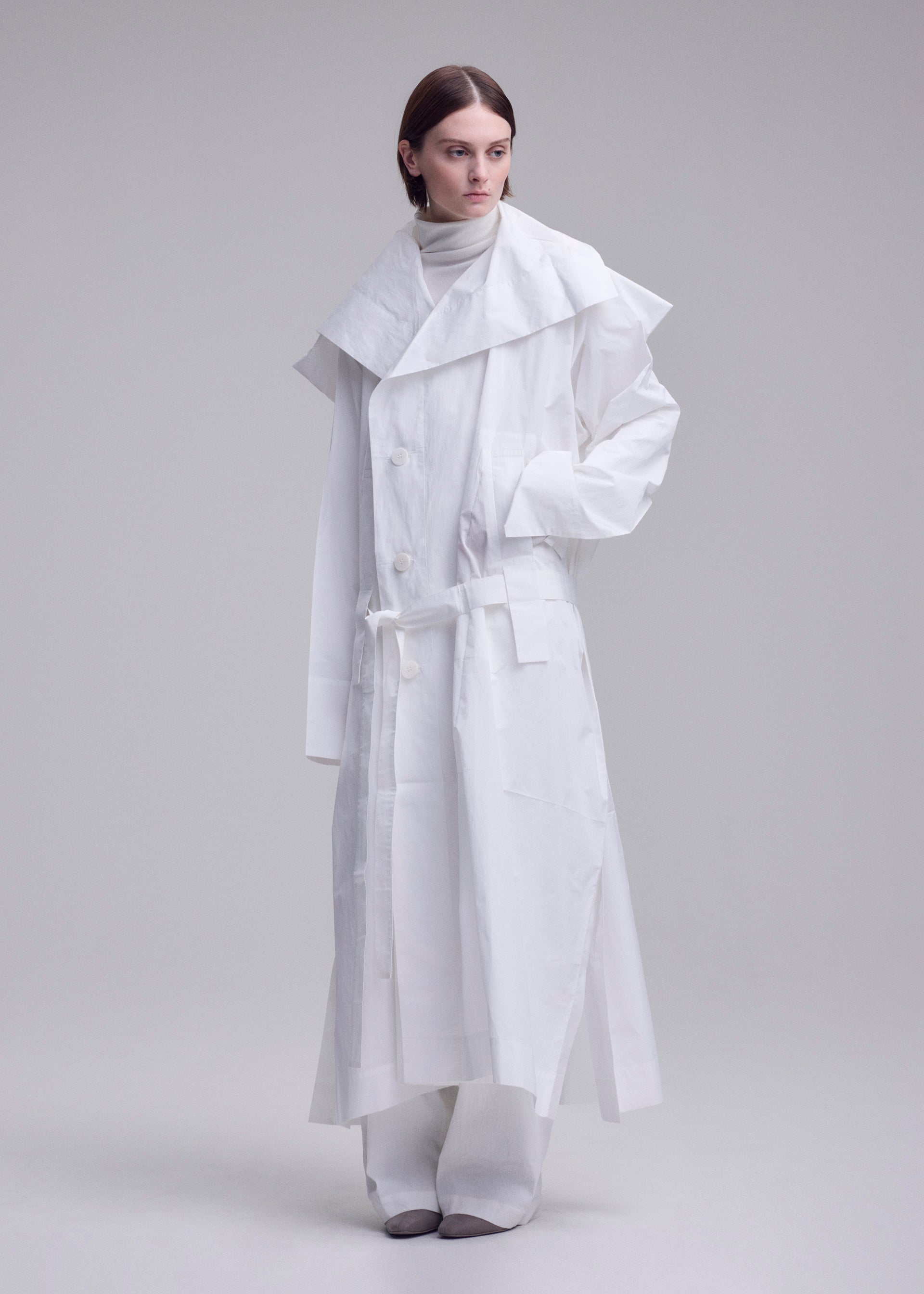 Zoomed view A person stands against a plain background wearing the ISSEY MIYAKE FOLDING COAT— a long white piece with a large collar, belted waist, and wide sleeves— layered over a high-neck top and wide-legged pants.