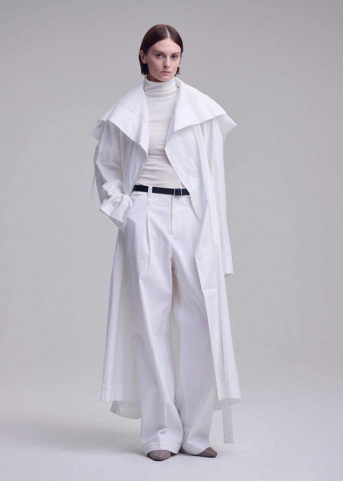 A person with short brown hair wears the FOLDING COAT by ISSEY MIYAKE over a white turtleneck and wide-leg trousers, styled with a black belt and gray shoes, standing against a plain light background. view 13 of 15