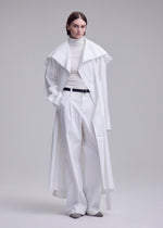 A person with short brown hair wears the FOLDING COAT by ISSEY MIYAKE over a white turtleneck and wide-leg trousers, styled with a black belt and gray shoes, standing against a plain light background.