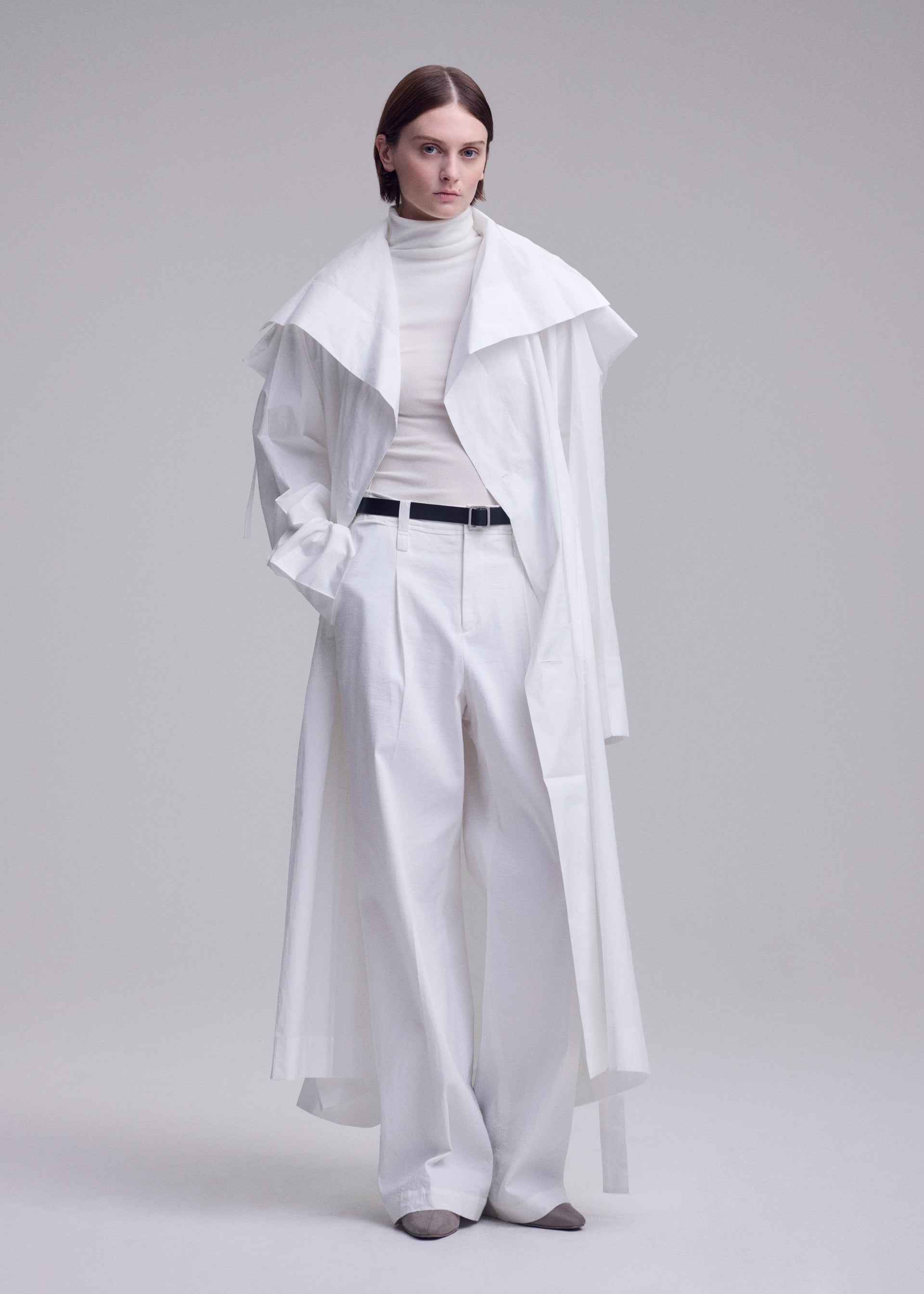 Zoomed view A person with short brown hair wears the FOLDING COAT by ISSEY MIYAKE over a white turtleneck and wide-leg trousers, styled with a black belt and gray shoes, standing against a plain light background.