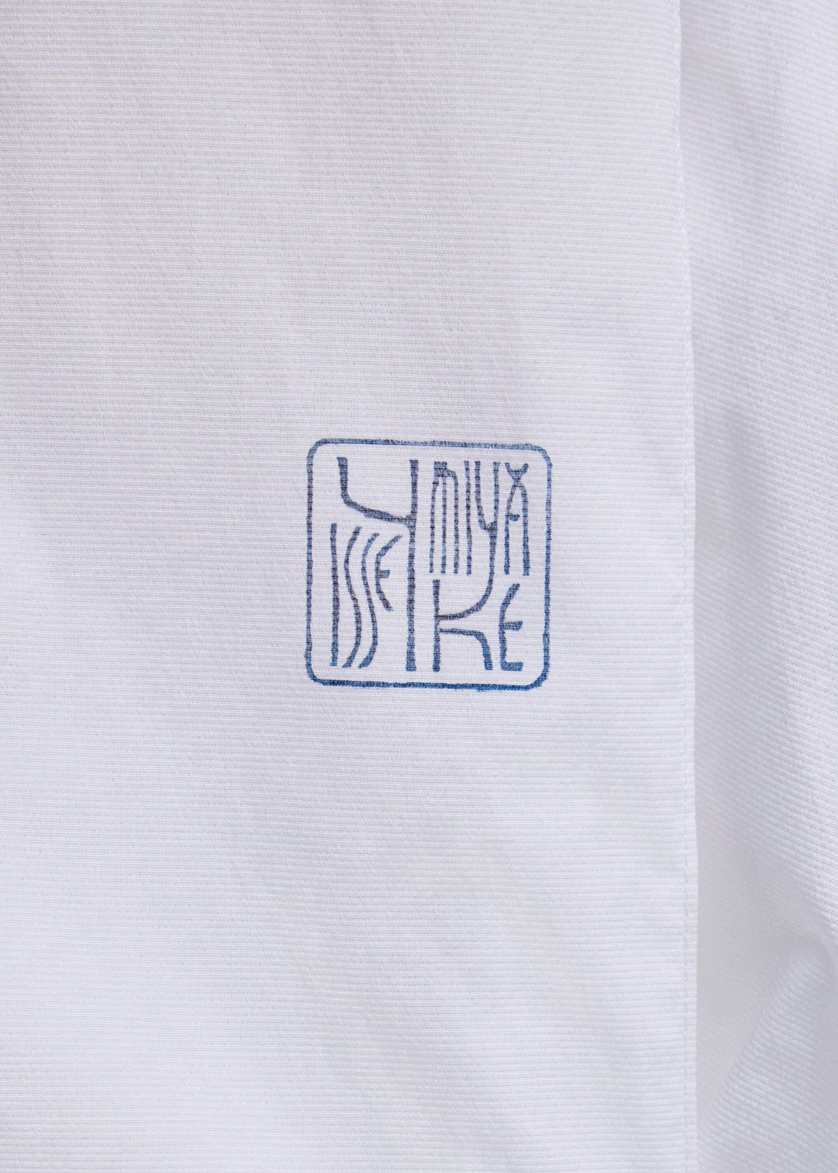 Close-up of white fabric from the ISSEY MIYAKE FOLDING COAT, featuring a blue, square-shaped stamp with ISSEY MIYAKE in stylized, abstract letters. view 5 of 15