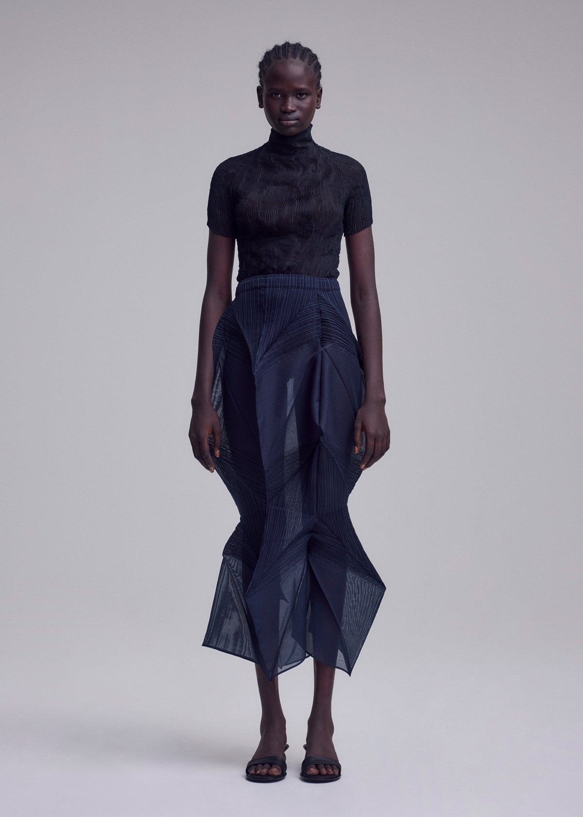A person stands against a plain background, wearing the ISSEY MIYAKE SHADE AND SHADED_NY black short-sleeved turtleneck top and sheer navy skirt, paired with black open-toe sandals. They have short braided hair and a neutral expression. view 4 of 11