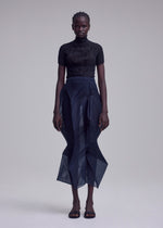 A person stands against a plain background, wearing the ISSEY MIYAKE SHADE AND SHADED_NY black short-sleeved turtleneck top and sheer navy skirt, paired with black open-toe sandals. They have short braided hair and a neutral expression.