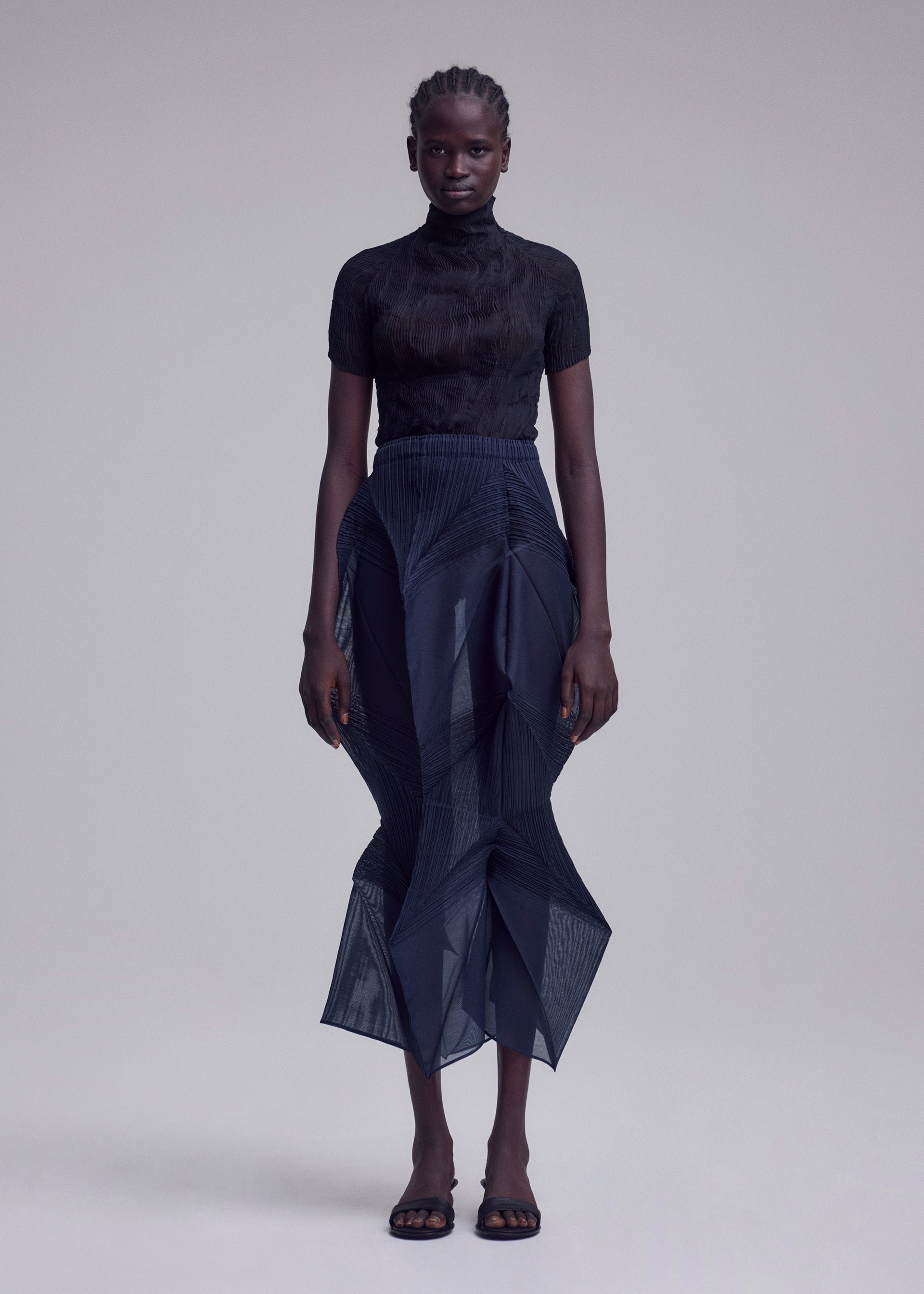 Zoomed view A person stands against a plain background, wearing the ISSEY MIYAKE SHADE AND SHADED_NY black short-sleeved turtleneck top and sheer navy skirt, paired with black open-toe sandals. They have short braided hair and a neutral expression.