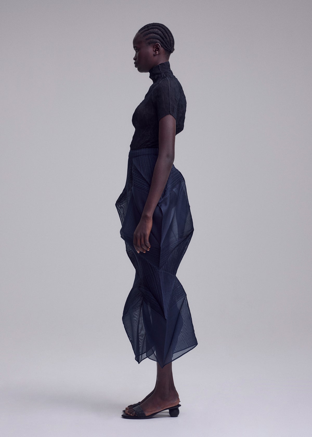 A person stands in profile against a plain background, wearing the ISSEY MIYAKE SHADE AND SHADED_NY sheer black short-sleeve top and sculptural semi-transparent navy skirt with black sandals. Their hair is styled in neat cornrows. view 6 of 11