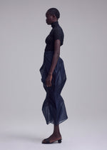 A person stands in profile against a plain background, wearing the ISSEY MIYAKE SHADE AND SHADED_NY sheer black short-sleeve top and sculptural semi-transparent navy skirt with black sandals. Their hair is styled in neat cornrows.