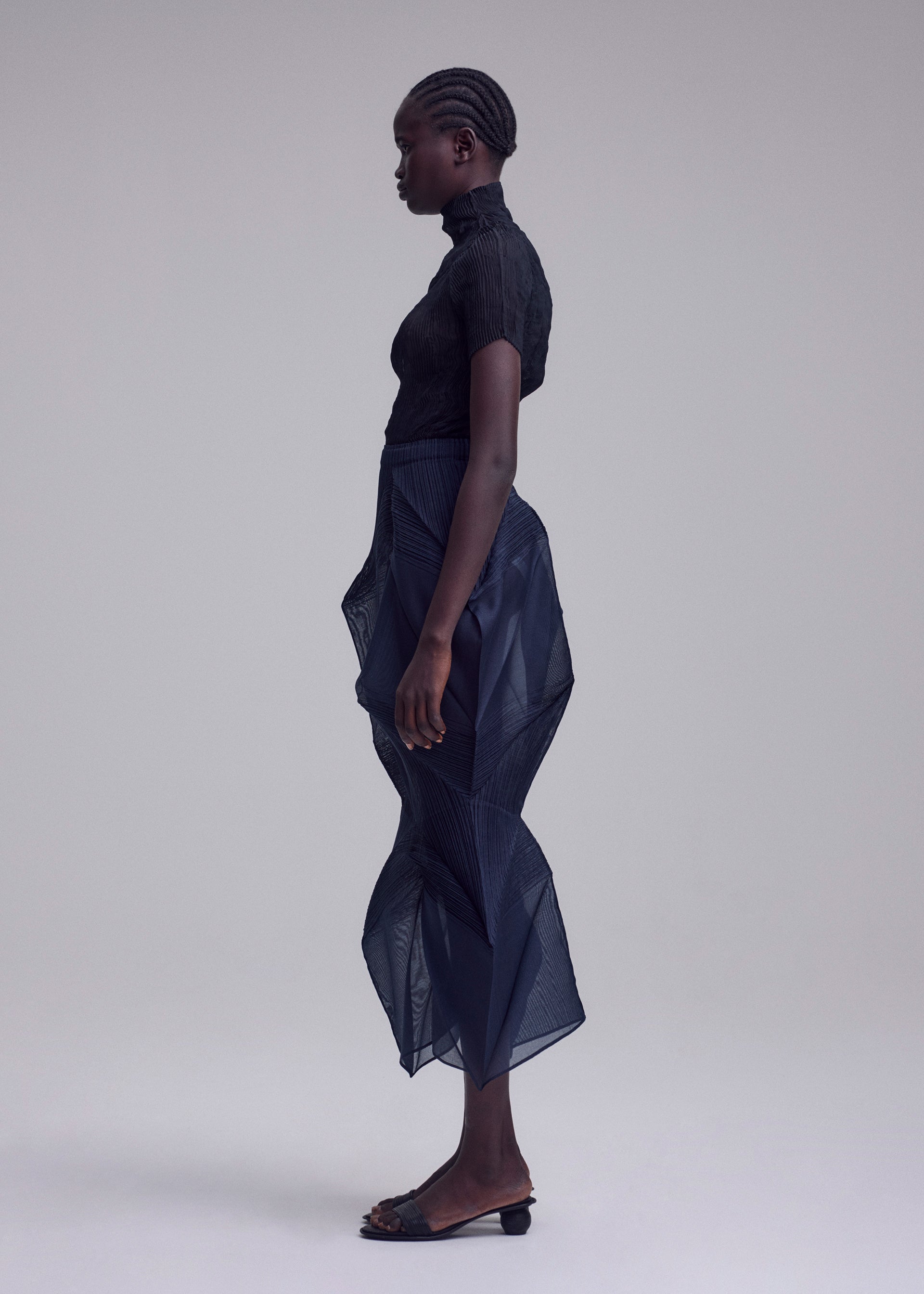 Zoomed view A person stands in profile against a plain background, wearing the ISSEY MIYAKE SHADE AND SHADED_NY sheer black short-sleeve top and sculptural semi-transparent navy skirt with black sandals. Their hair is styled in neat cornrows.