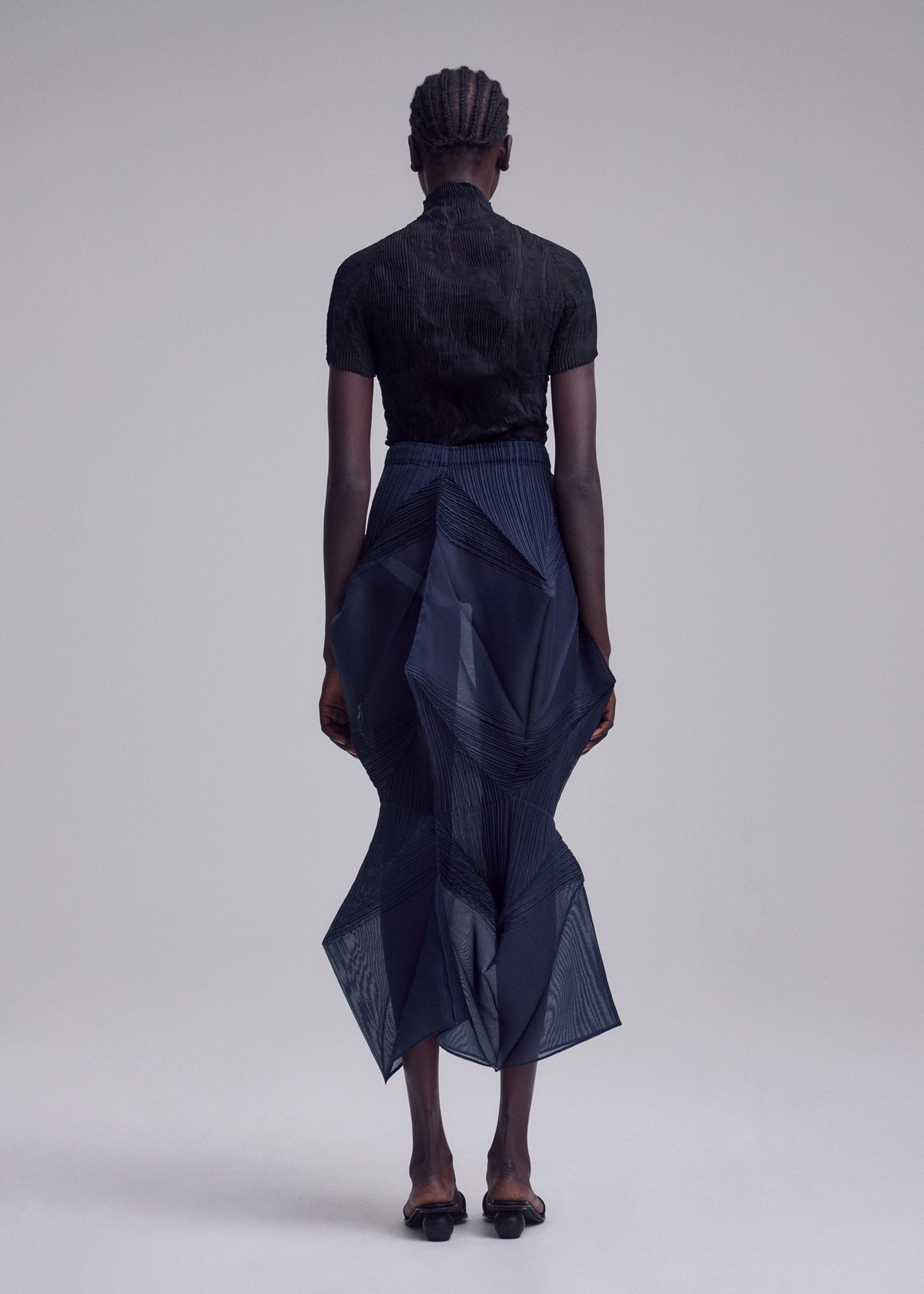 A person with braided hair faces away, wearing ISSEY MIYAKE's SHADE AND SHADED_NY: a black short-sleeved top with a dark sheer, geometric skirt featuring an asymmetrical hem and black sandals, set against a plain light background. view 7 of 11