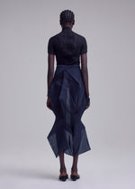 A person with braided hair faces away, wearing ISSEY MIYAKE's SHADE AND SHADED_NY: a black short-sleeved top with a dark sheer, geometric skirt featuring an asymmetrical hem and black sandals, set against a plain light background.
