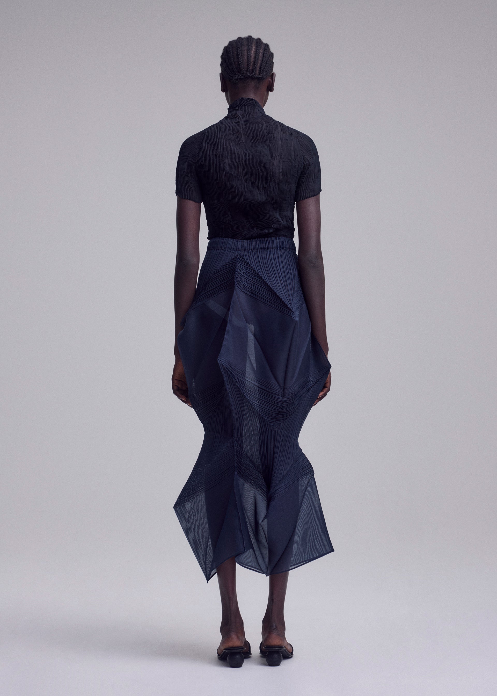 Zoomed view A person with braided hair faces away, wearing ISSEY MIYAKE's SHADE AND SHADED_NY: a black short-sleeved top with a dark sheer, geometric skirt featuring an asymmetrical hem and black sandals, set against a plain light background.