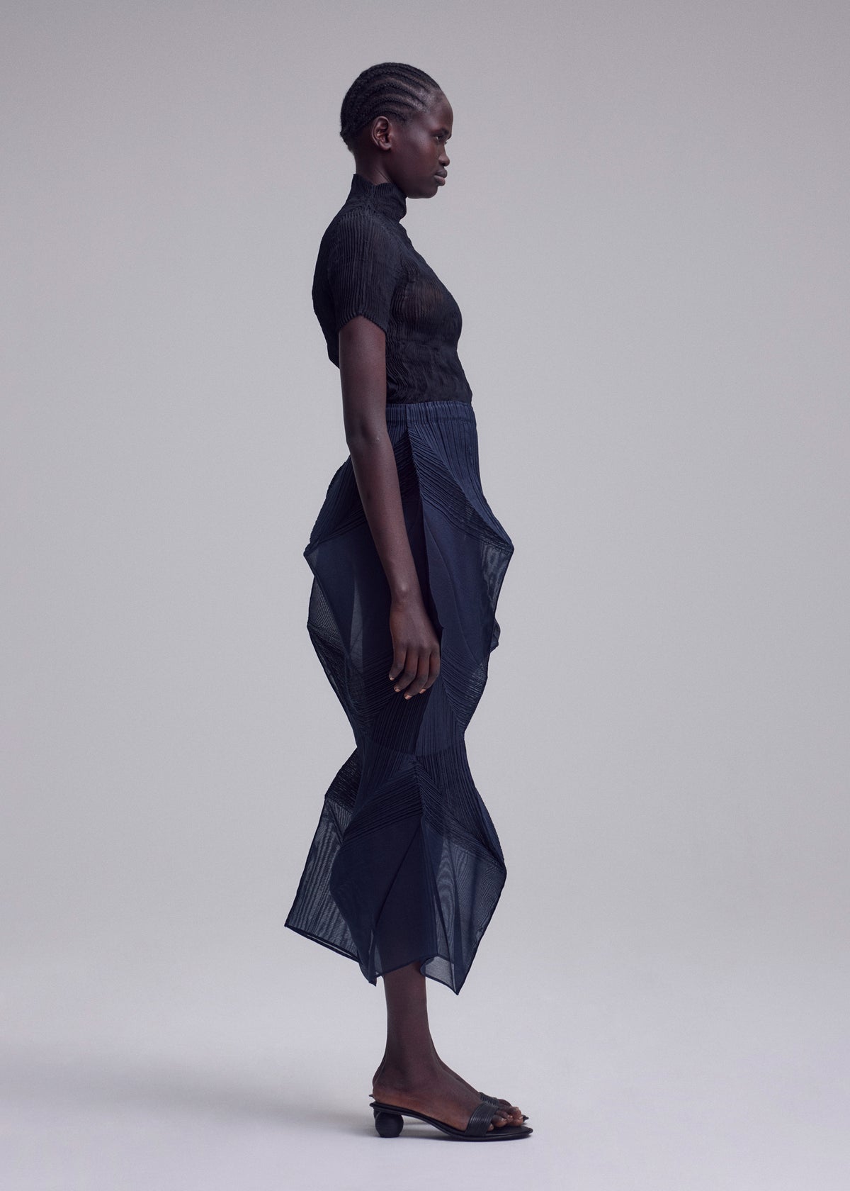 A person stands in profile against a plain background, wearing the SHADE AND SHADED_NY top and skirt by ISSEY MIYAKE, paired with open-toed black low-heeled sandals. view 8 of 11