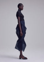 A person stands in profile against a plain background, wearing the SHADE AND SHADED_NY top and skirt by ISSEY MIYAKE, paired with open-toed black low-heeled sandals.