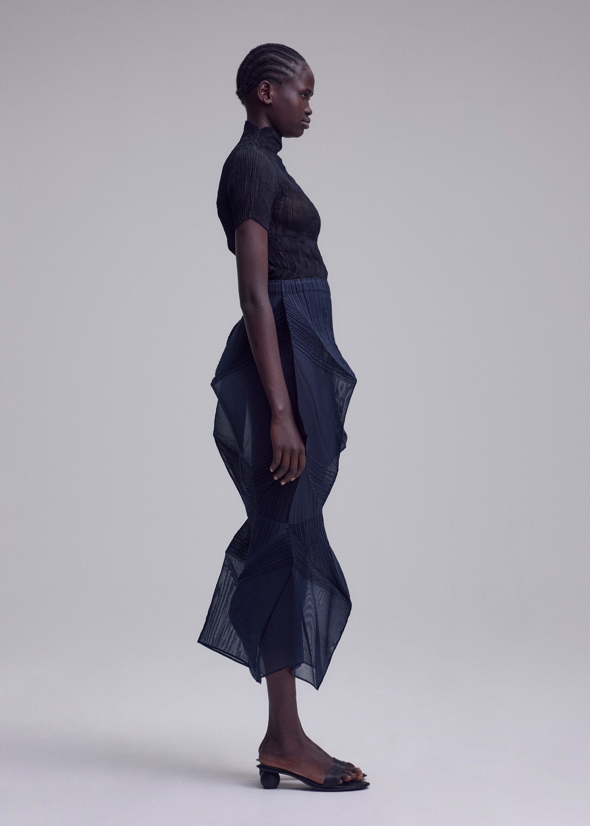 Zoomed view A person stands in profile against a plain background, wearing the SHADE AND SHADED_NY top and skirt by ISSEY MIYAKE, paired with open-toed black low-heeled sandals.