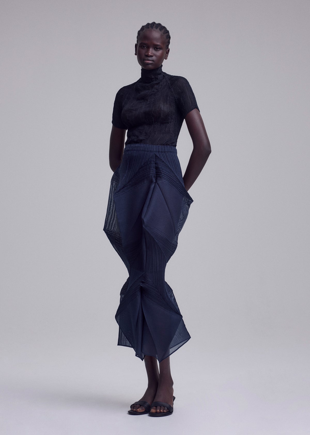 A person stands against a plain backdrop, wearing ISSEY MIYAKE's SHADE AND SHADED_NY: a fitted black short-sleeve top, navy sculptural pleated skirt, and black sandals. Their hands are behind their back and their expression is neutral. view 5 of 11