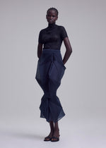 A person stands against a plain backdrop, wearing ISSEY MIYAKE's SHADE AND SHADED_NY: a fitted black short-sleeve top, navy sculptural pleated skirt, and black sandals. Their hands are behind their back and their expression is neutral.