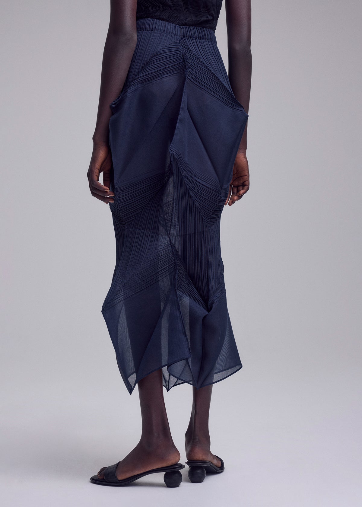 A person stands against a plain background, wearing the SHADE AND SHADED_NY sheer dark blue geometric skirt by ISSEY MIYAKE and black open-toe sandals with rounded heels. Their arms are relaxed at their sides. view 10 of 11
