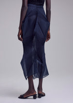 A person stands against a plain background, wearing the SHADE AND SHADED_NY sheer dark blue geometric skirt by ISSEY MIYAKE and black open-toe sandals with rounded heels. Their arms are relaxed at their sides.