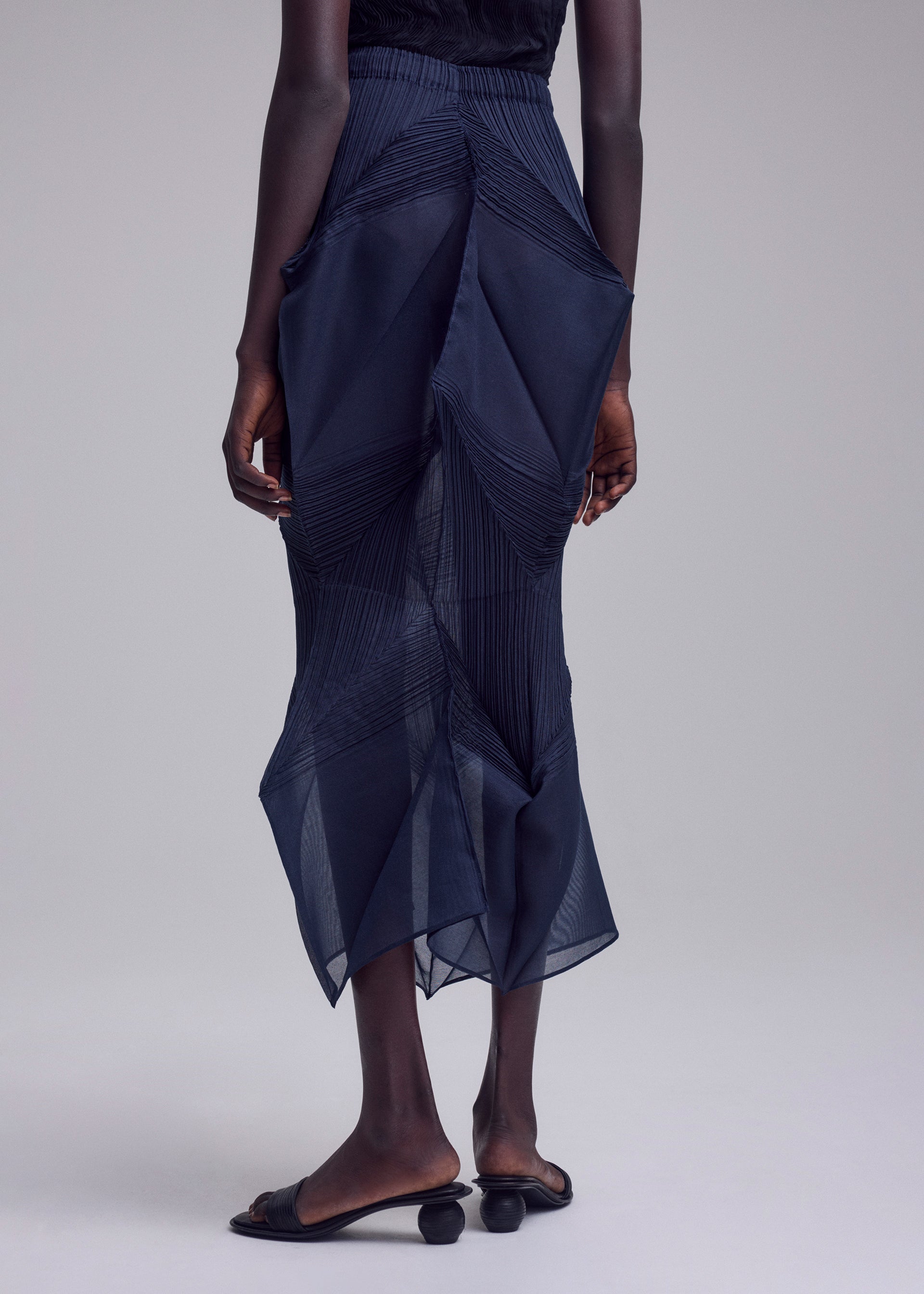 Zoomed view A person stands against a plain background, wearing the SHADE AND SHADED_NY sheer dark blue geometric skirt by ISSEY MIYAKE and black open-toe sandals with rounded heels. Their arms are relaxed at their sides.