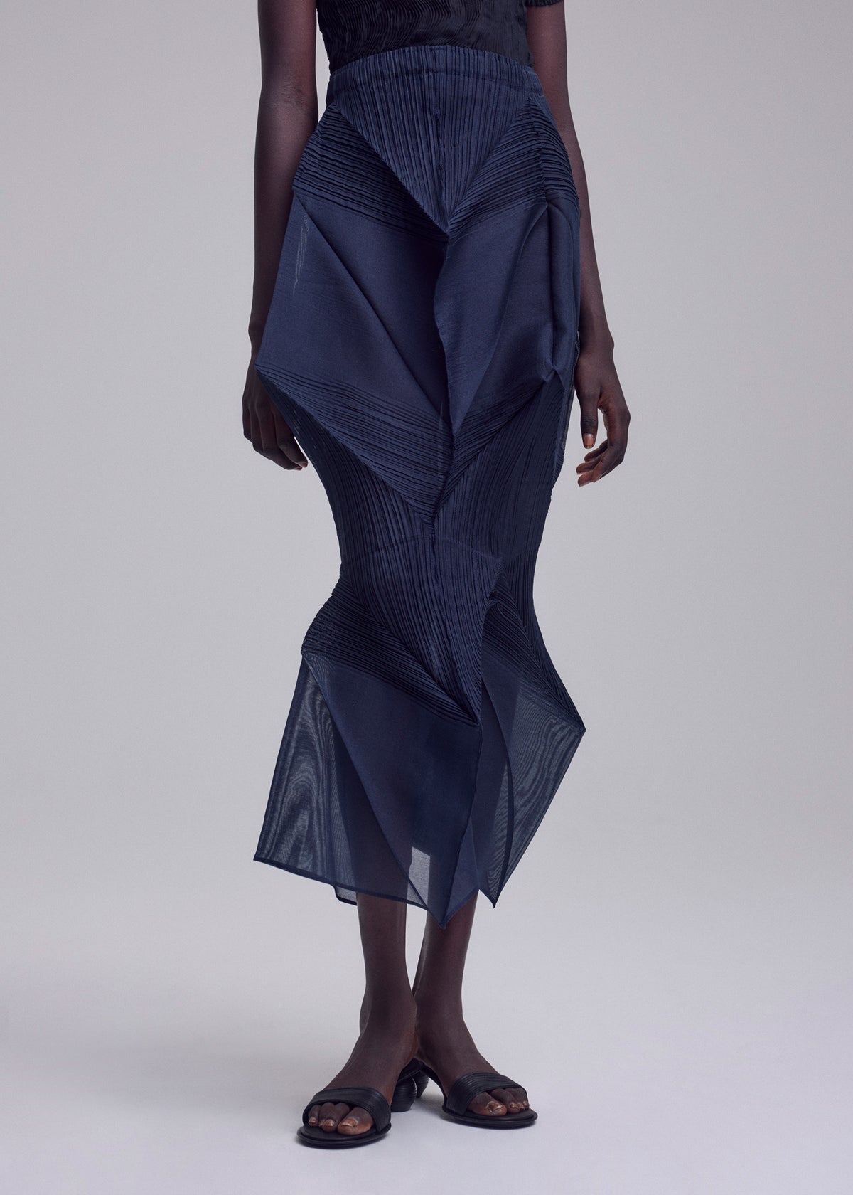 A person wears the SHADE AND SHADED_NY skirt by ISSEY MIYAKE—an architectural dark pleated skirt with an asymmetrical hem and sheer panels—paired with black slide sandals against a plain light background. view 9 of 11