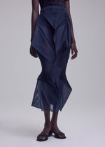 A person wears the SHADE AND SHADED_NY skirt by ISSEY MIYAKE—an architectural dark pleated skirt with an asymmetrical hem and sheer panels—paired with black slide sandals against a plain light background.