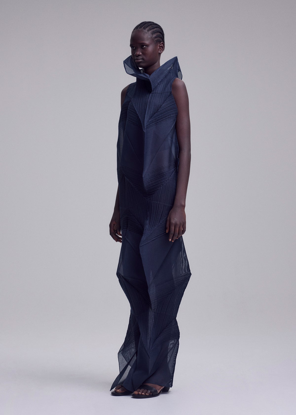 A person with short, braided hair wears the SHADE AND SHADED_NY long dress by ISSEY MIYAKE, featuring a dark geometric design and sculptural silhouette, standing against a plain light gray background. view 4 of 10