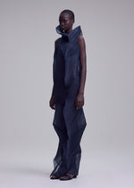 A person with short, braided hair wears the SHADE AND SHADED_NY long dress by ISSEY MIYAKE, featuring a dark geometric design and sculptural silhouette, standing against a plain light gray background.