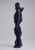 A person in profile wears ISSEY MIYAKE's SHADE AND SHADED_NY, a futuristic long dress with machine-pleated, geometric shapes and a high collar. The sculptural silhouette stands out against the plain light background.