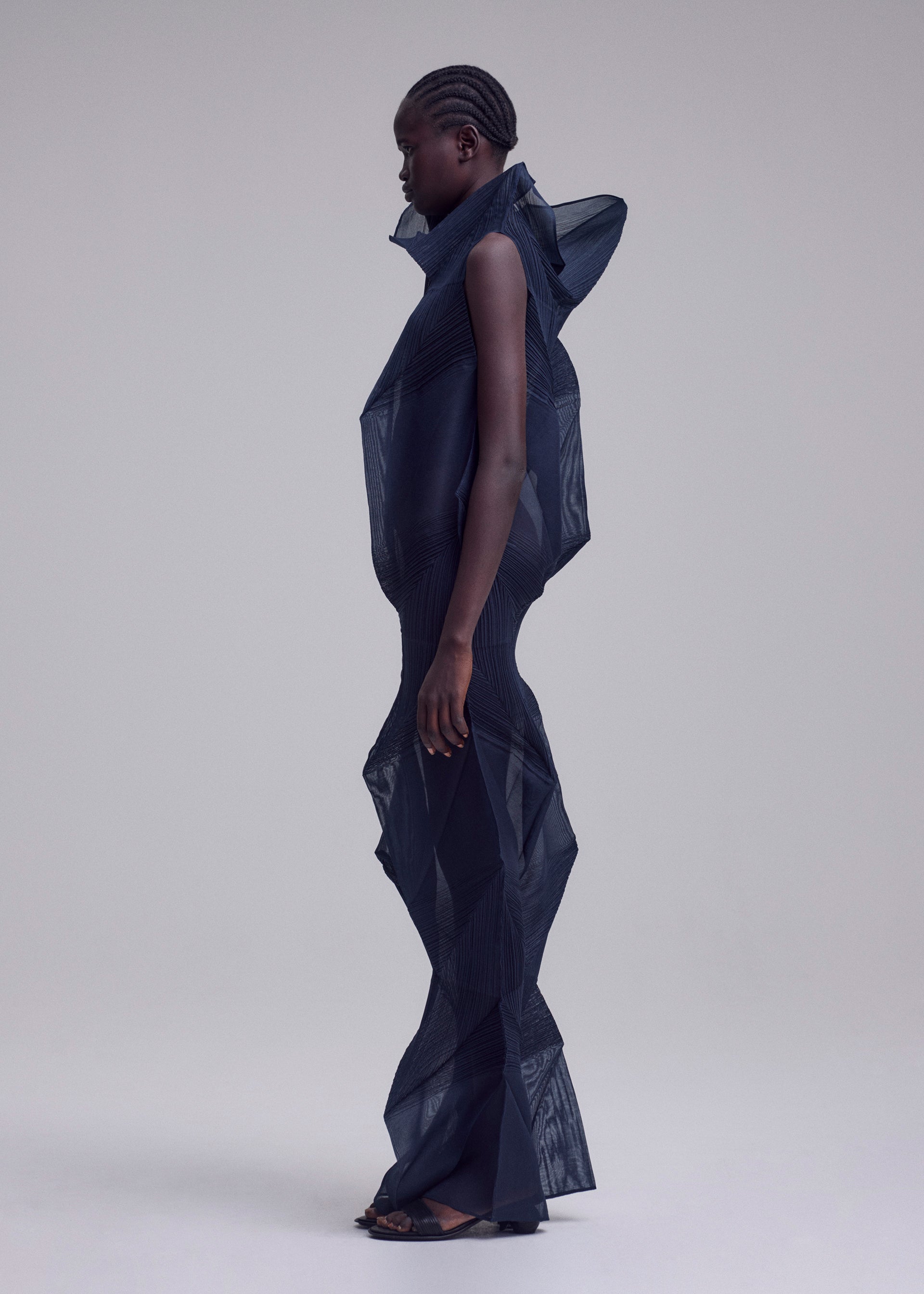 Zoomed view A person in profile wears ISSEY MIYAKE's SHADE AND SHADED_NY, a futuristic long dress with machine-pleated, geometric shapes and a high collar. The sculptural silhouette stands out against the plain light background.
