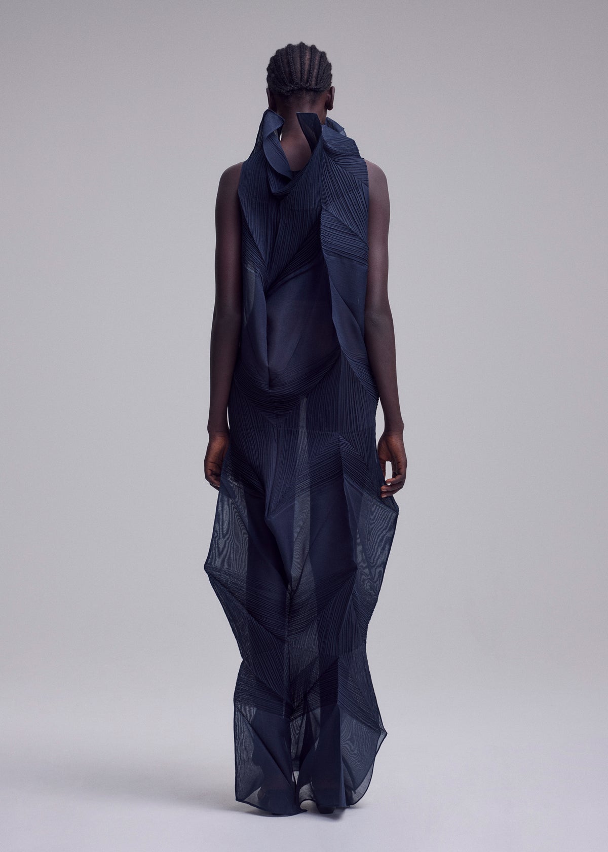 With short, braided hair and facing away, the person wears ISSEY MIYAKE’s SHADE AND SHADED_NY: a sheer dark blue long dress with geometric patterns and layered, sculptural fabric, set against a plain light gray background. view 6 of 10